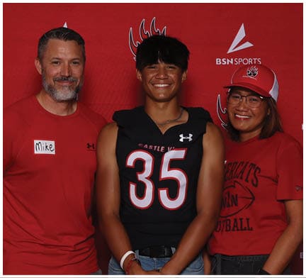Three people stand smiling in front of a red backdrop with sports logos, with one person wearing a sports jersey and the others wearing red shirts. Description generated by AI
