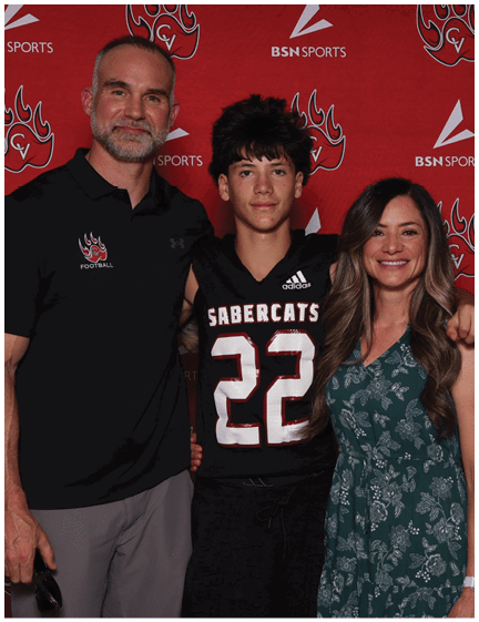 Three people stand closely together smiling in front of a red backdrop, with one person wearing a sports jersey numbered 22 and the others dressed casually. Description generated by AI