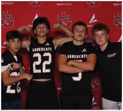 Two people wearing black Sabercats sports jerseys pose confidently with one flexing muscles and the other crossing arms against a red backdrop. Description generated by AI