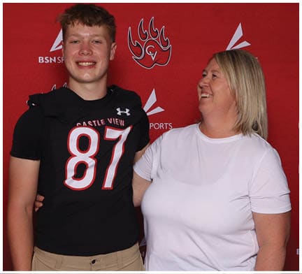 A person wearing a black sports jersey with the number 87 stands smiling next to another person in a white shirt who is looking at them and smiling against a red backdrop with logos. Description generated by AI
