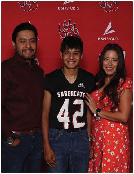 Three people stand closely together smiling in front of a red backdrop with sports logos, with one person wearing a black sports jersey and the others dressed in casual and floral clothing. Description generated by AI