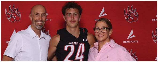 Three people stand together smiling in front of a red backdrop with logos, with one person wearing a black sports jersey numbered 24 and the others wearing casual button-up shirts. Description generated by AI