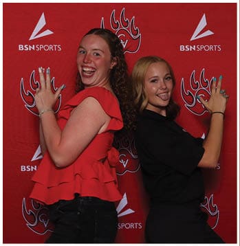 Two adults stand back to back smiling and posing playfully in front of a red backdrop with sports logos. Description generated by AI