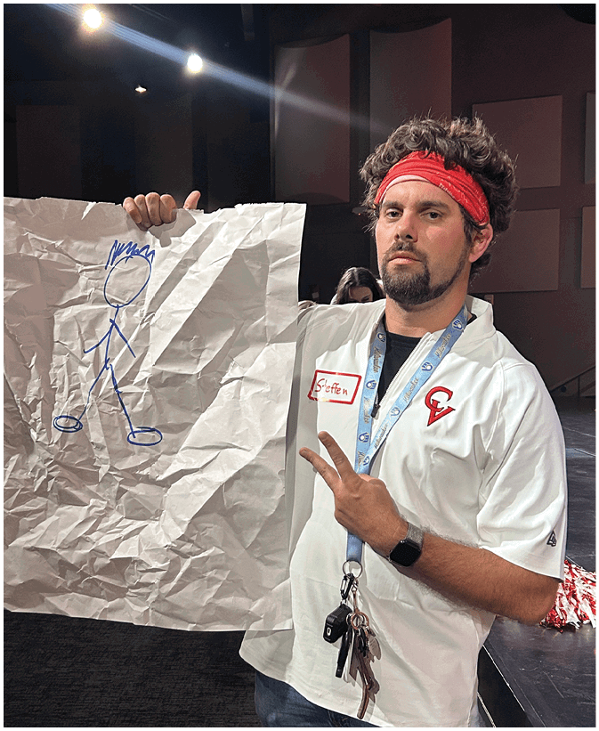 A person wearing a white shirt with a red headband holds a large crumpled paper with a blue stick figure drawing while making a peace sign with the other hand. Description generated by AI