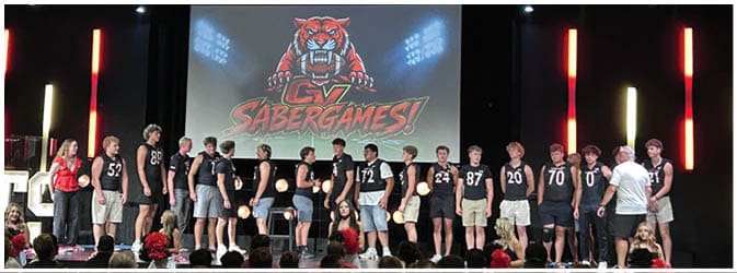 A stage with a large screen displaying a fierce tiger mascot logo is illuminated by vertical red lights and overhead spotlights in a dark room. Description generated by AI