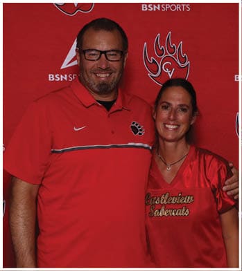 A person wearing glasses and a red polo shirt with a paw print logo is smiling in front of a red backdrop with sports logos. Description generated by AI