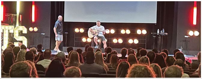 Two people are on a stage with one sitting and playing a guitar while the other stands nearby, with a large audience watching and illuminated vertical lights and a sign that reads “TATS" in the background. Description generated by AI