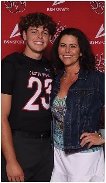 Two people stand closely together smiling in front of a red backdrop with sports logos, one wearing a black sports jersey with the number 23 and the other wearing a denim jacket over a patterned top and white skirt. Description generated by AI