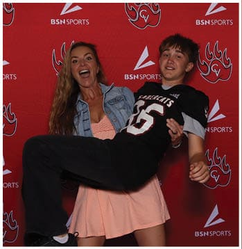 A person wearing a denim jacket is holding another person dressed in a black sports jersey with the number 66 in front of a red backdrop with sports logos. Description generated by AI