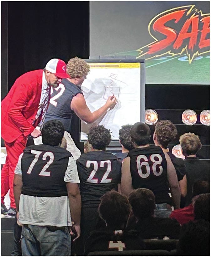 Two adults are engaged in drawing on a large pad of paper on an easel in front of an audience with a colorful graphic projected on a screen behind them. Description generated by AI