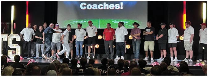 A group of adults standing on a stage in casual clothing with a large audience in front and a screen behind them displaying the word “Coaches!" Description generated by AI