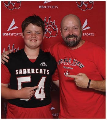 Two people smiling and posing together in front of a red backdrop with sports logos, one wearing a black sports jersey with the number 54 and the other wearing a red shirt with text. Description generated by AI