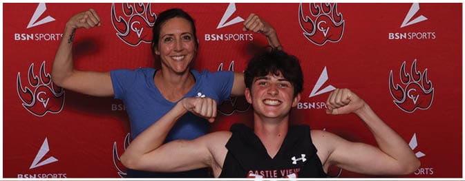 A person smiling and flexing both arms in front of a red backdrop with repeated logos of a stylized paw print and the text BSN SPORTS. Description generated by AI
