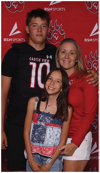 Three people stand closely together smiling in front of a red backdrop with sports logos, one wearing a black sports jersey, another in a red long-sleeve shirt and white shorts, and the third in a patterned sleeveless top and denim shorts. Description generated by AI