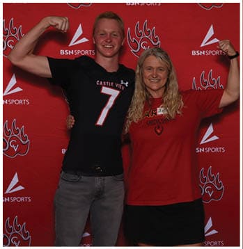 Two people smiling and flexing their arms in front of a red backdrop with sports logos, one wearing a black sports jersey with the number seven and the other wearing a red t-shirt and black skirt. Description generated by AI