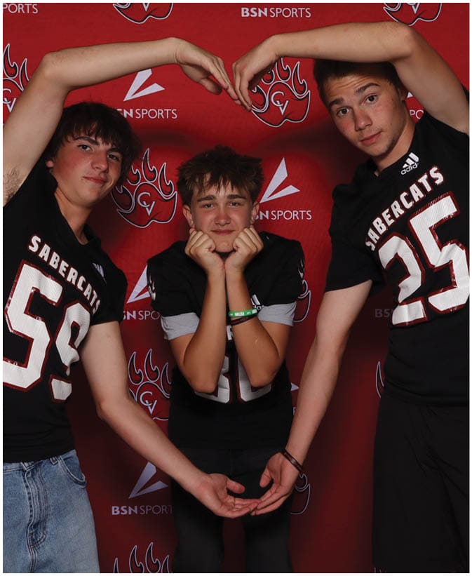 Three people wearing black sports jerseys pose in front of a red backdrop with logos, with two forming a heart shape with their arms around the third person who is smiling and resting their chin on their hands. Description generated by AI