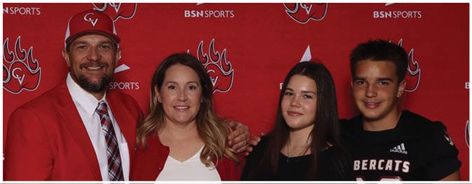 Four people stand closely together smiling in front of a red backdrop with sports logos, with one person wearing a red jacket and cap. Description generated by AI