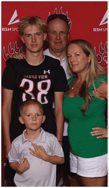 A group of four people, including two adults and two children, pose closely together in front of a red backdrop with sports logos, with one child wearing a black sports shirt and the other wearing a light-colored polo. Description generated by AI