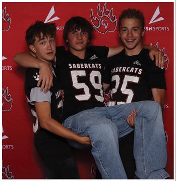 Three people wearing black Sabercats sports jerseys pose smiling against a red backdrop, with two holding the third person who is wearing jeans and sneakers. Description generated by AI