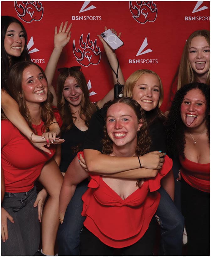 Five people smiling and posing playfully in front of a red backdrop with sports logos, with two people giving piggyback rides. Description generated by AI