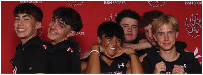 Five people wearing black sports jerseys are standing closely together in front of a red backdrop, smiling and posing playfully. Description generated by AI