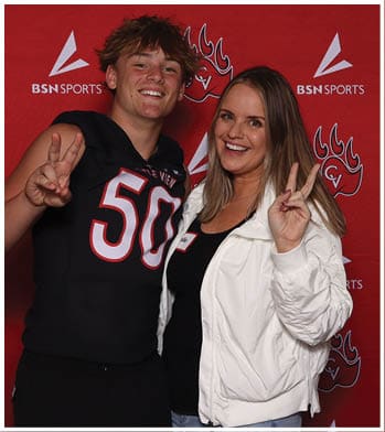 Two people stand in front of a red backdrop with sports logos, one wearing a black sports jersey and the other wearing a white jacket and making a peace sign while smiling. Description generated by AI
