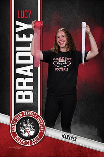 A person with curly hair wearing a black Castle View Football t-shirt is smiling and holding up two rolls of athletic tape, one red and one white, standing in front of a backdrop with the name Lucy Bradley and Castle View Varsity Football Class of 2021. Description generated by AI