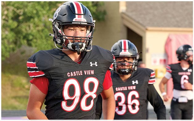 Two people wearing black football uniforms with red and white stripes and helmets are on a field, with one person in the foreground wearing number 98 and the other in the background wearing number 36. Description generated by AI