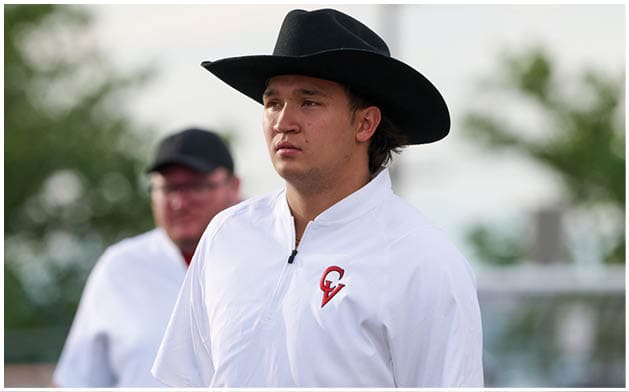 A person wearing a black cowboy hat and a white shirt with a red and black logo stands outdoors with another person in the background. Description generated by AI