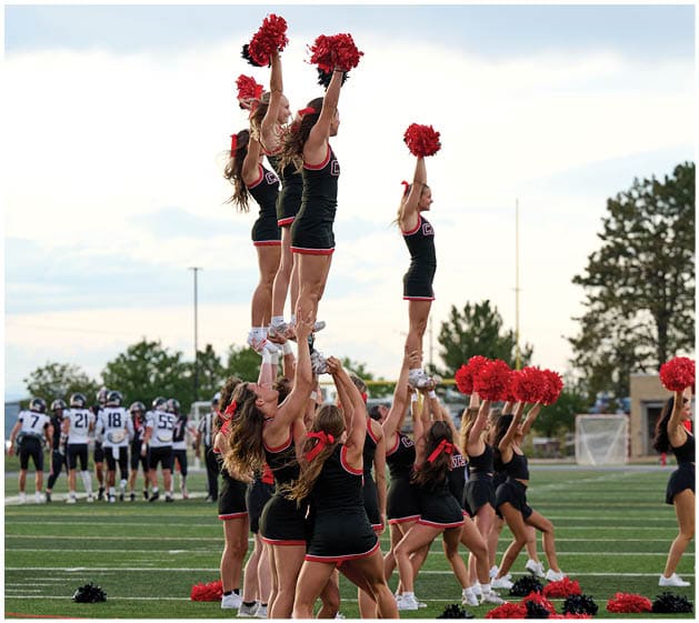 A group of cheerleaders in black and red uniforms perform a stunt on a football field while football players stand in the background. Description generated by AI