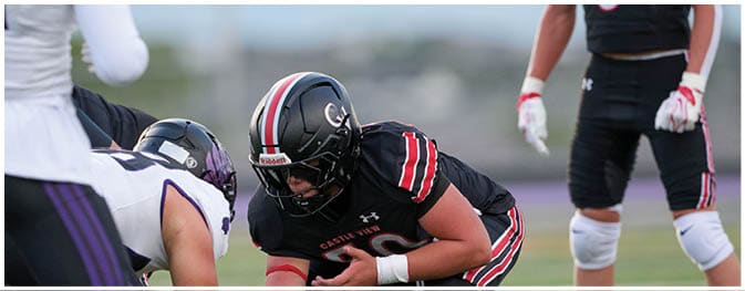 People in football uniforms are positioned on a field preparing for a play during a game. Description generated by AI