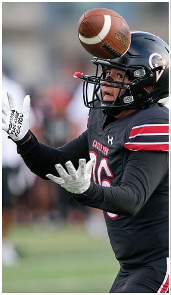 A person wearing a black football helmet and jersey with the number 16 is focused on catching a football in mid-air. Description generated by AI