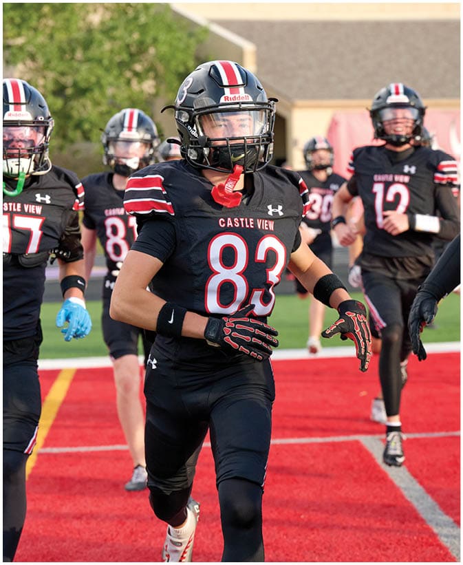 People in black football uniforms with red and white stripes are running on a red field, with some wearing helmets and gloves. Description generated by AI