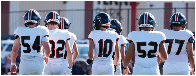 Five people wearing football helmets and white jerseys with numbers stand on a field facing away from the camera. Description generated by AI