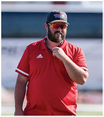 A person wearing a red Adidas polo shirt, a black cap with “SABERCATS FOOTBALL" text, and red reflective sunglasses is standing outdoors. Description generated by AI