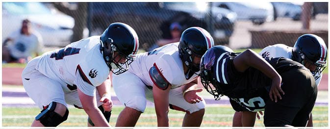 Three people in football uniforms and helmets are crouched on a field facing each other, preparing for a play. Description generated by AI