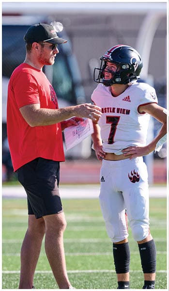 An adult wearing a red shirt and cap is talking to a person in a football uniform with a helmet on a sports field. Description generated by AI