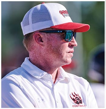 A person wearing a white and red baseball cap with “SABER FOOTBALL" embroidered on it and dark sunglasses is looking to the right outdoors. Description generated by AI