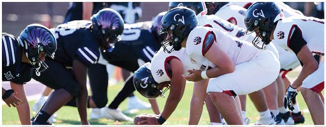 Two opposing American football teams in helmets and uniforms are lined up facing each other on a grassy field, preparing for a play. Description generated by AI