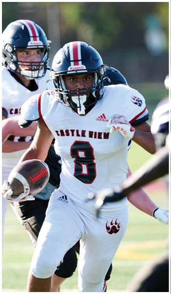 A person wearing a white football uniform with “Castle View" and the number 8 is running with a football while being pursued by opponents on a field. Description generated by AI