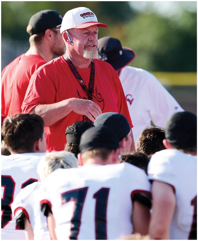 A group of people wearing sports jerseys listens attentively to two adults in red shirts and hats who appear to be coaches giving instructions outdoors. Description generated by AI