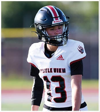 A person wearing a black helmet and a white football jersey with the number 13 and the words “Castle View" stands on a field looking to the side. Description generated by AI