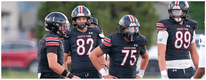 Four people wearing black football uniforms with helmets stand together on a field during a game or practice. Description generated by AI