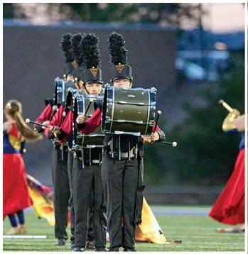 A group of people in marching band uniforms play drums in formation on a field while others in colorful costumes perform with flags in the background. Description generated by AI