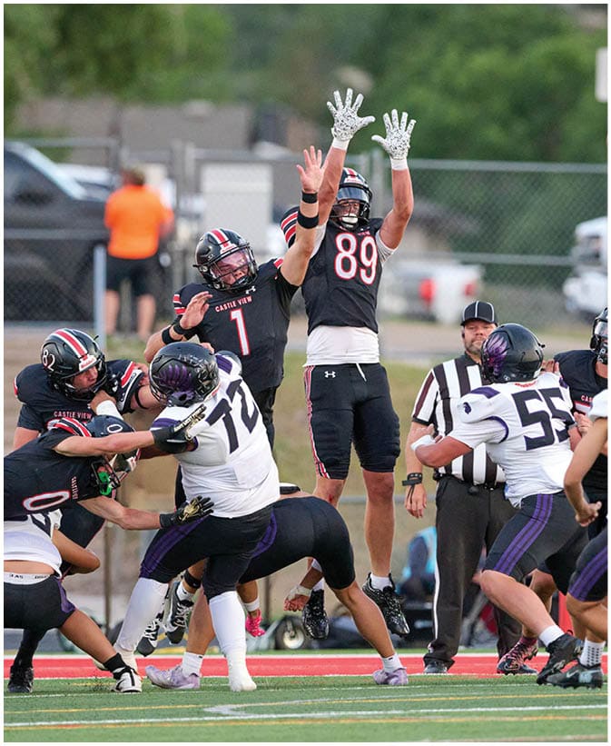 Several football players in black and white uniforms are actively engaged in a play on a field with one player in black jumping with arms raised and others attempting to block or tackle. Description generated by AI