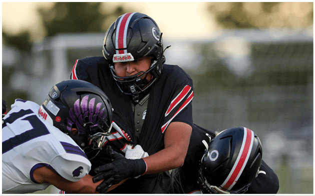 Three people wearing football helmets and uniforms are engaged in a close physical play on a football field. Description generated by AI