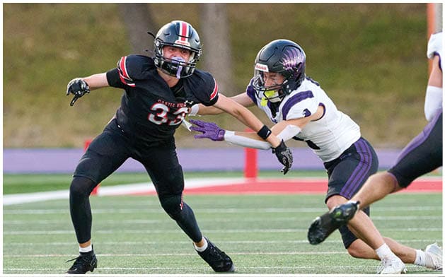 Two people in football uniforms are engaged in a play on a field with one person reaching out to block or tackle the other. Description generated by AI