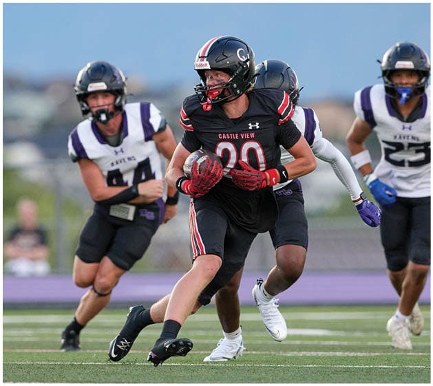 A person wearing a black football uniform with the number 20 is running with the ball while being pursued by three people in white and purple uniforms on a football field. Description generated by AI