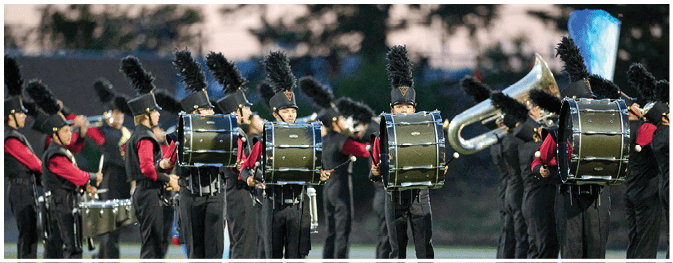 A marching band dressed in black and red uniforms with tall black plumes on their hats is performing outdoors during dusk. Description generated by AI