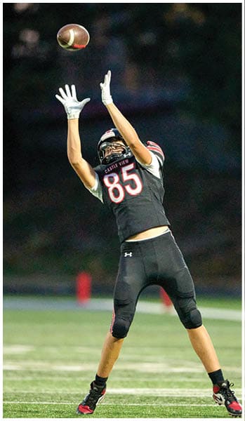 A person in a black football uniform with the number 85 is reaching up to catch a football while another person in casual clothing stands nearby on a football field. Description generated by AI